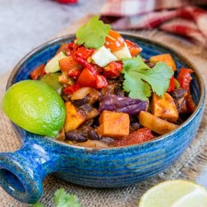 A close up of a blue bowl full of Brazilian roasted vegetable feijoada. A wedge of lime and leaves of coriander garnish the dish.
