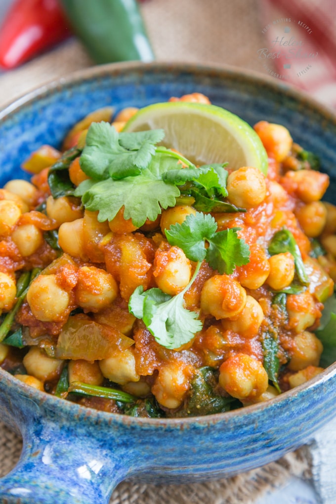 A close up of a blue bowl holding an individual serving of Indian chickpea curry with tomato and spinach. The dish is garnished with a wedge of lime and some leaves of fresh coriander.