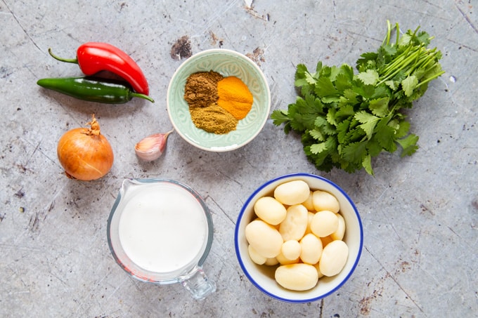 A top down view of ingredients for potato curry with a bowl of spices, chillies, coconut milk, coriander and canned potatoes