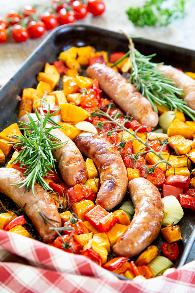 Colourful red tomatoes and peppers, yellow butternut squash and sausages cooked until brown, garnished with fresh green rosemary.