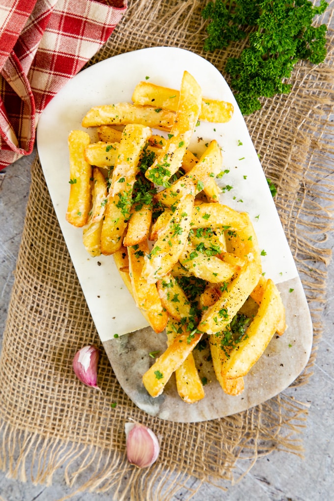 A top down picture of golden garlic Parmesan fries on a marble board, garnished with parsley.