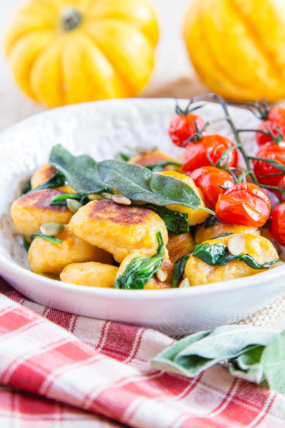 A bowl of golden pumpkin gnocchi with crispy fried sage and cherry tomatoes on the side, set on a red and white cloth with winter squash in the background.