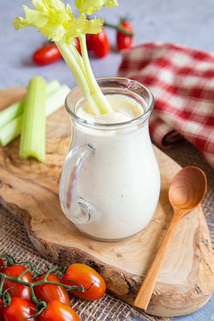 A stick of celery sticks out of a jug of salad cream. Around the jug are tomatoes, a napkin & spoon, and more celery.