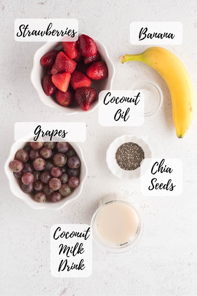 The ingredients for strawberry coconut smoothie; an annotated picture taken from above, listing strawberries, banana, coconut oil, chia seeds, grapes, and coconut milk drink.