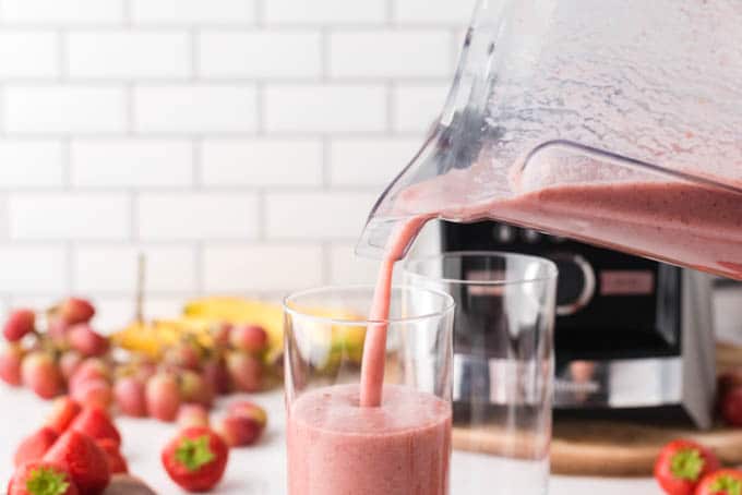 A thick pink smoothie is being poured out of a blender jug into a glass. Out of focus in the background are the blender itself and more fruit.