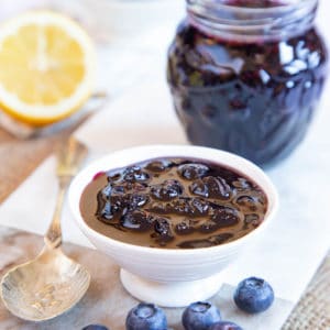 A close up of a small dish of blueberry jam. A pot of the same is in the background, as is half a lemon.