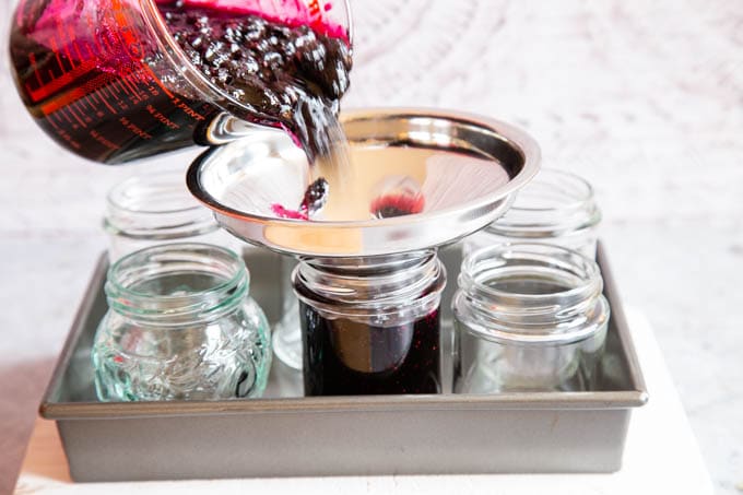 Filling jars with jam using a glass jug and jam funnel. The jars are arranged in a roasting tin.