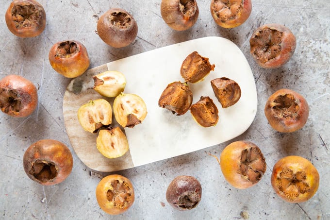 Brown bletted and pale unbletted medlars cut open on a chopping board, surrounded by more fruit.
