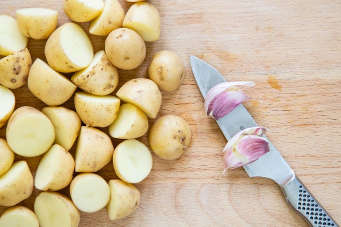 A wooden chopping board half covered with halved baby potatoes, with a knife and 2 cloves of garlic on the other half.
