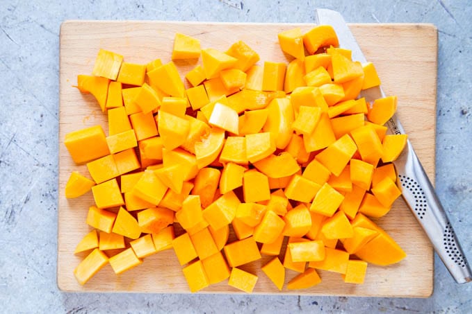 Cubed orange butternut squash on a wooden chopping board. A large kitchen knife is under some cubes.
