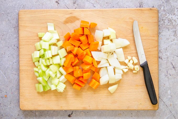 A wooden board showing chopped onions, carrots and celery. A black handled knife is also on the board.