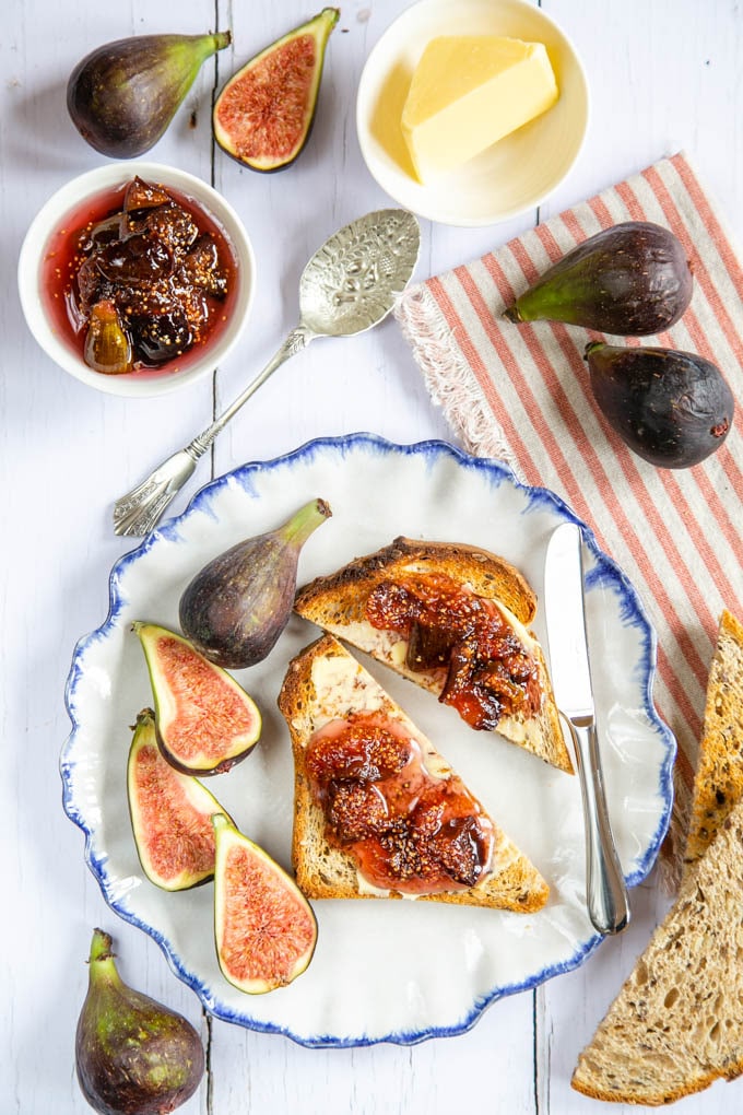 A top down picture of a small bowl of jam, next to a plate with toast spread with butter and jam. Figs sit next to the plate.