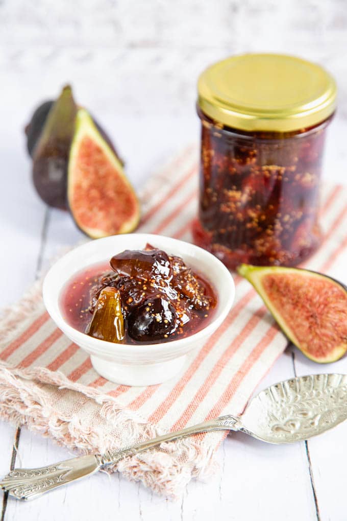 Close up picture of a small bowl of fig jam, with a full jar in the background. There are also whole and halved figs surrounding the bowl.