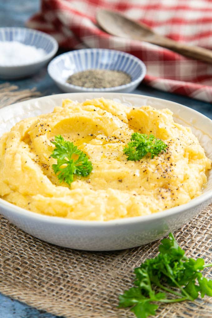 A close up of a bowl of heaped golden yellow swede mash, garnished with parsley leaves and sprinkled with black pepper.