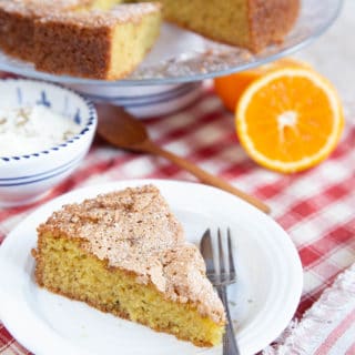 Close up of a slice of orange olive oil cake. Half an orange and the rest of the cake is out of focus in the background.