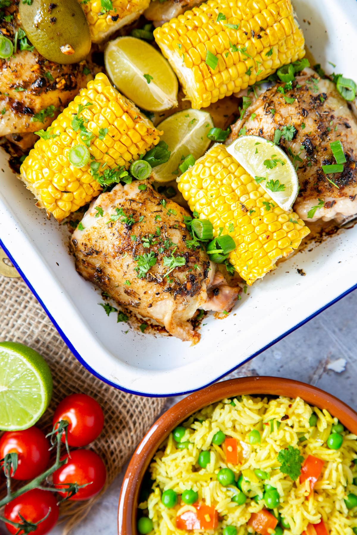 A close up of a roasting dish of cooked chicken thighs and corn on the cob. At the bottom of the frame is a dish of fragrant rice and peas.