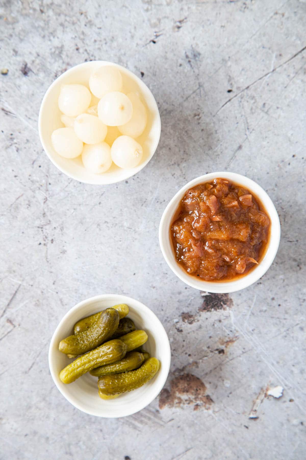 A top down picture of three small dishes holding pickled onions, chutney and pickled gherkins.