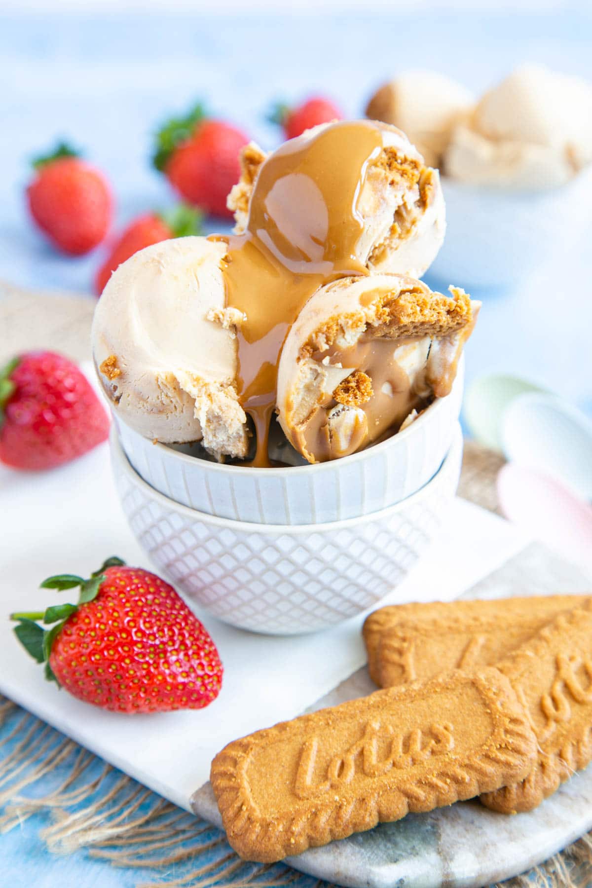 Three scoops of Biscoff ice cream in a stack of two bowls. In the foreground are some lotus speculoos biscuits and a strawberry. Melted speculoos has been poured onto the ice cream.