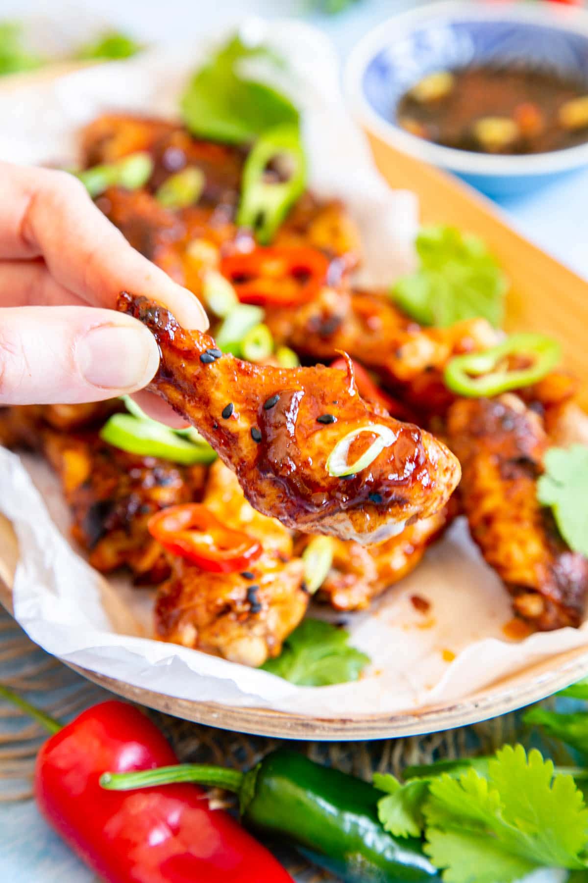 A hand holds a sticky Asian chicken wing above a wooden serving dish filled with more wings.