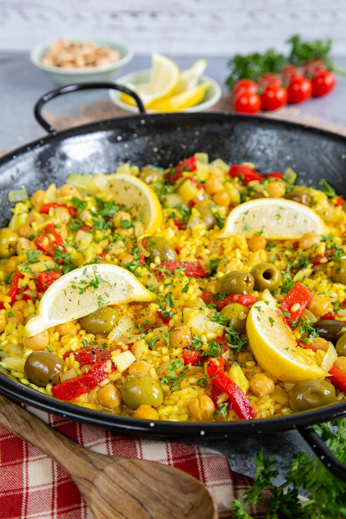 A close up on a black pan full of veggie paella, with lemon wedges on top. In the foreground is parsley, a cloth and a wooden spoon.