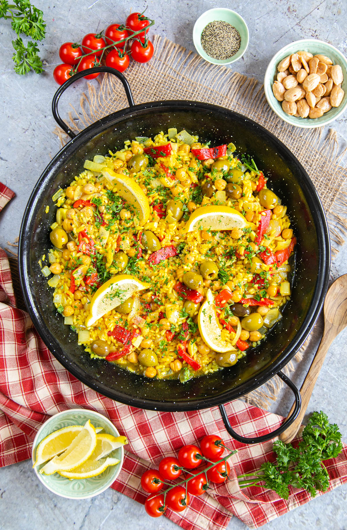 A top down of yellow easy vegetable paella, garnished with parsley and four wedges of lemon. Surrounding the pan are tomatoes on the vine, cloths, a wooden spoon, and dishes of almonds, pepper, and lemon wedges.