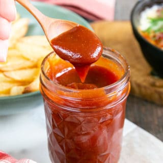Close up of glossy red barbecue sauce on a small wooden spoon, above a jar holding more sauce.