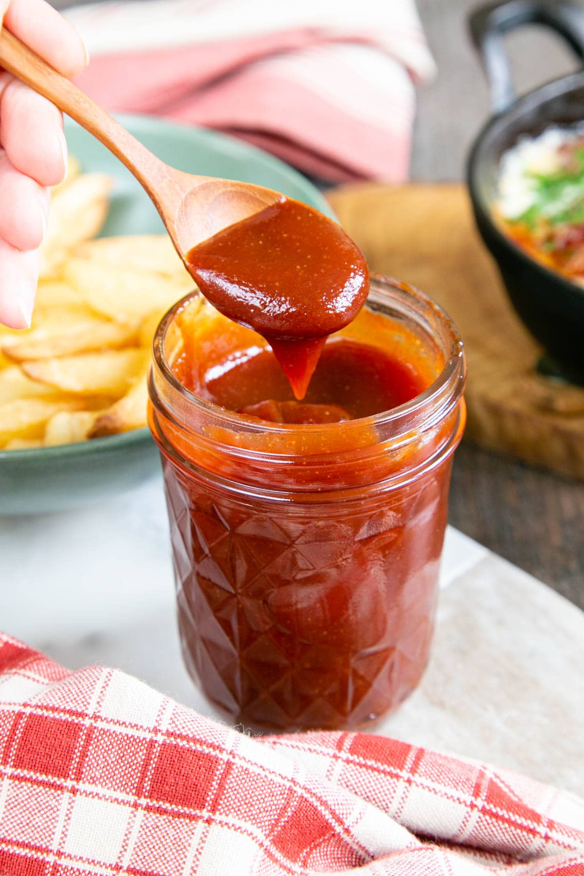 A jeweled faceted jam jar full of delicious dark red BBQ sauce. A small wooden spoon has spooned out a dollop, holding it above the jar.