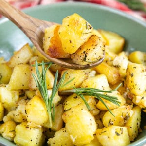 Close up of parmentier potatoes, seasoned with ground black pepper and sprigs of rosemary. A wooden spoon is held above the potatoes, holding a three cubes of potato.