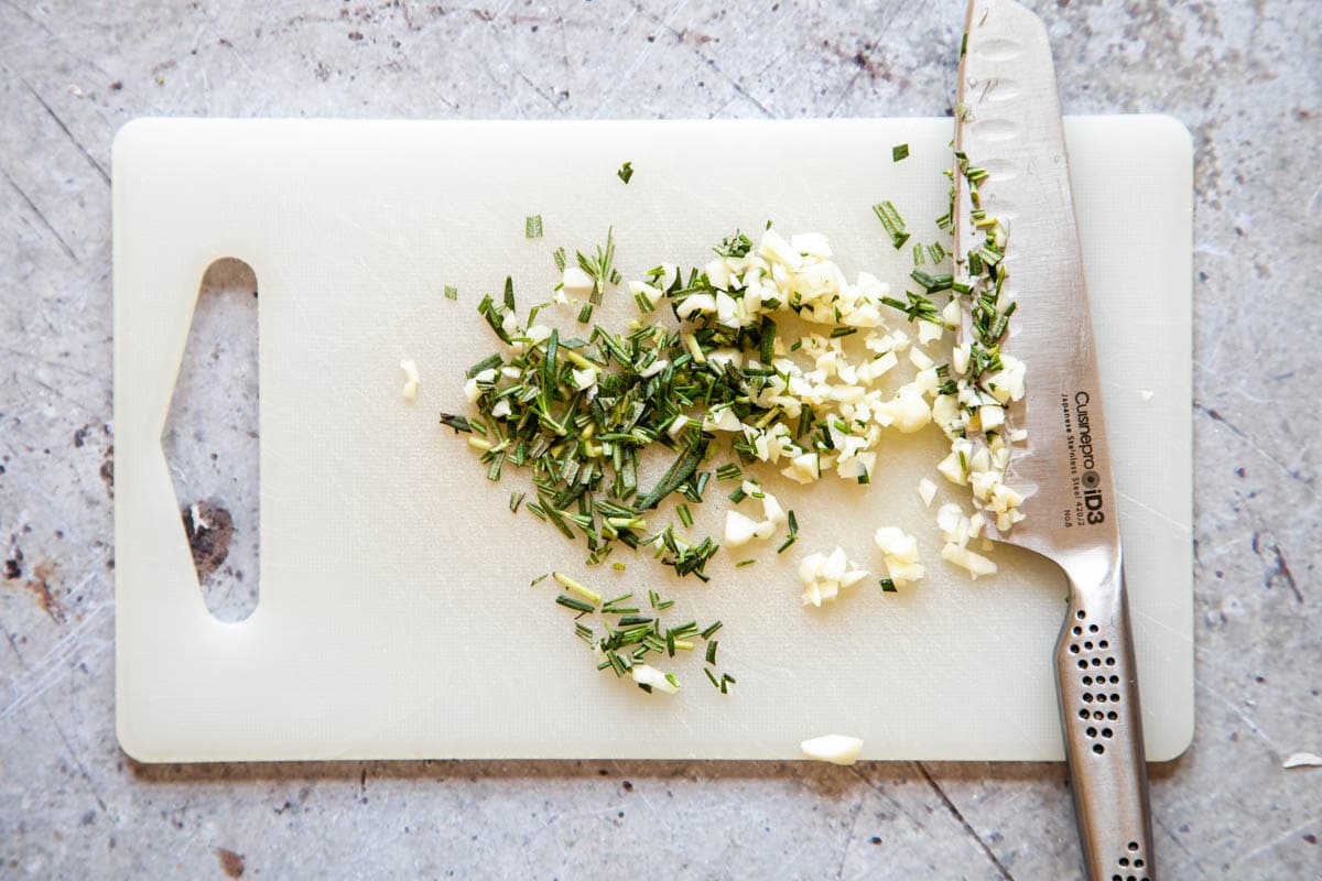 A white chopping board with chopped rosemary leaves and garlic.