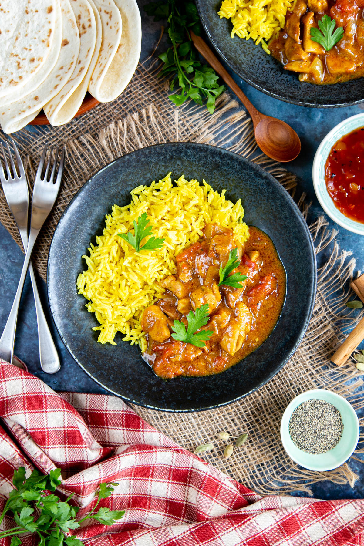 Delicious slow cooker curry in a black bowl, served with yellow pilau rice. The curry has been garnished with coriander leaves, and is served with flatbreads and a small dish of mango chutney. Spices, cloths, forks surround the bowl.