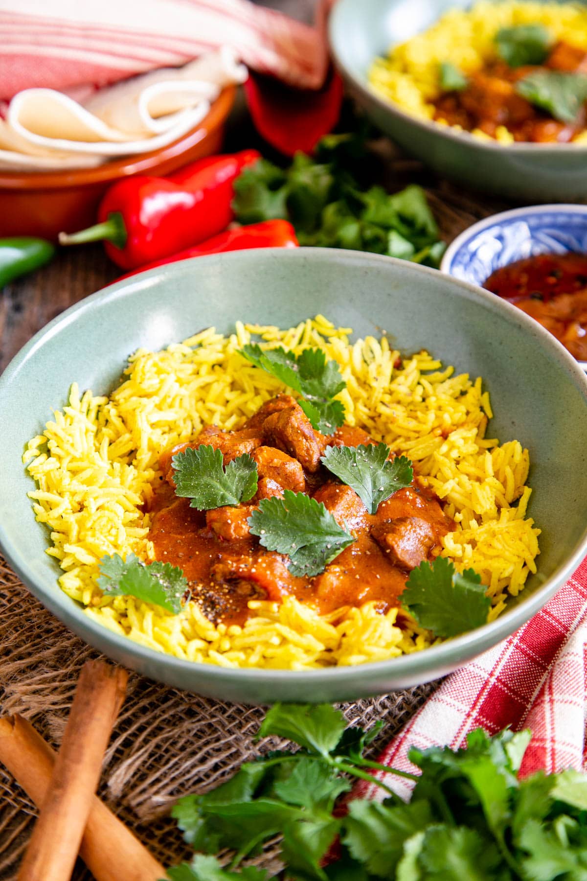 Slow cooker lamb curry, served in a green bowl with bright yellow pilau rice, garnished with coriander leaves. More coriander, cinnamon sticks, flatbreads, and chutney surround the bowl.