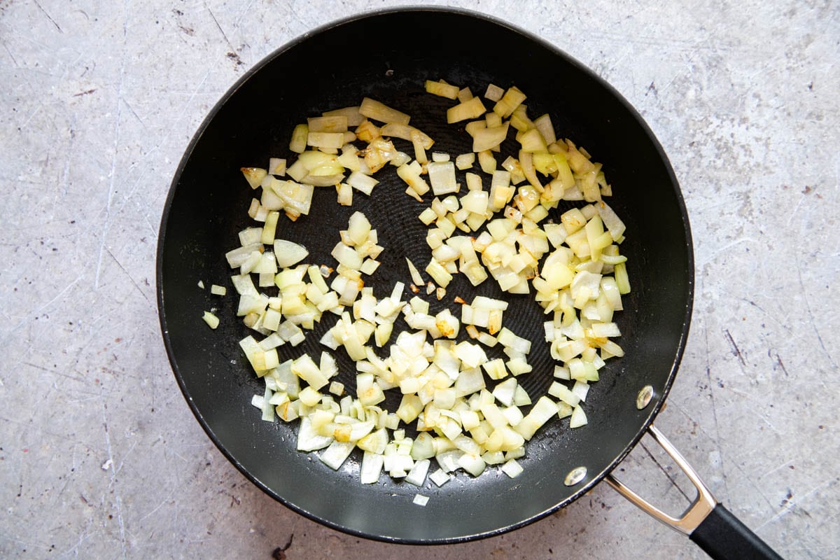 Diced onions frying in a little oil in a black frying pan. The onions are just starting to turn translucent.