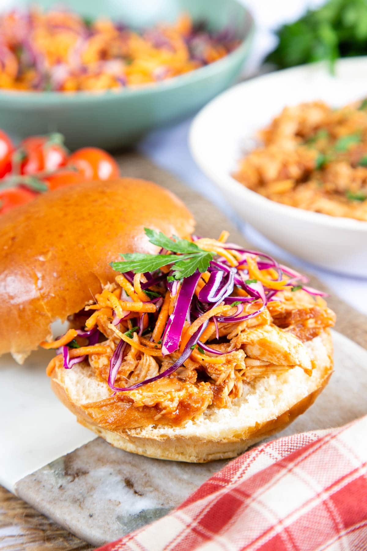 A close up of a burger bun piled with delicious BBQ slow cooked chicken, topped with red cabbage and carrot coleslaw. The bun is presented on a marble board.