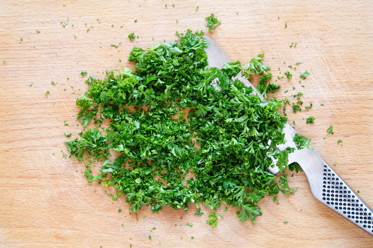 Bright green parsley being chopped on a wooden chopping board with a large kitchen knife.