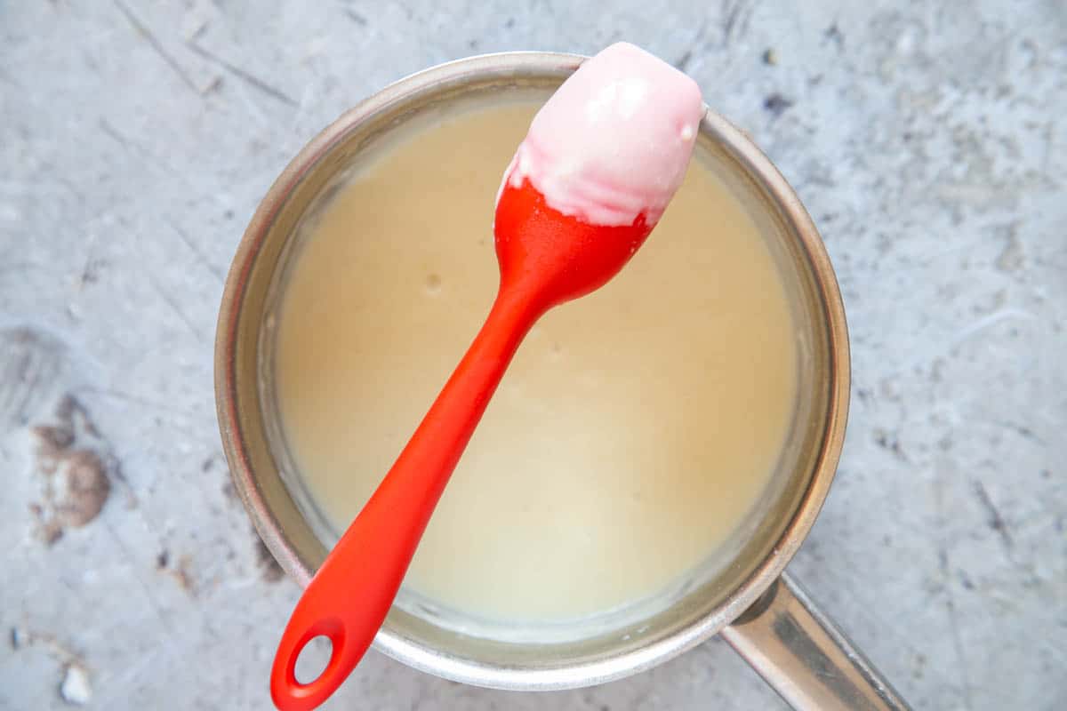 White sauce evenly covers the back of a red silicone spoon, balanced on a saucepan full of more sauce.