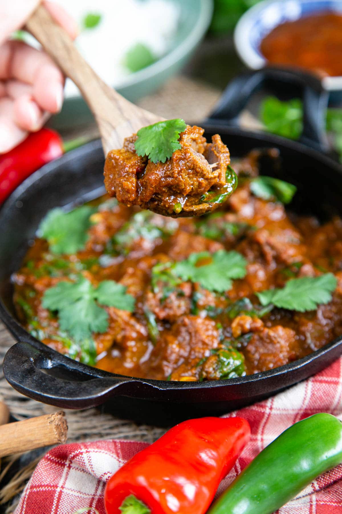 A spoonful of luscious, glossy slow cooker beef curry being served in a wooden spoon; the curry has been taken from a black iron serving dish in the background of the shot. Red and green chillies, and a red and white gingham cloth are in the foreground.