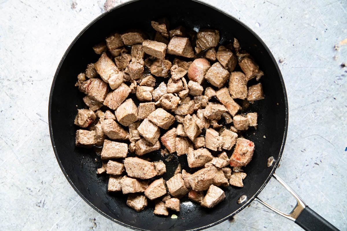 Cubes of beef being fried in a black frying pan. The beef has been just cooked enough to colour the outside of the cubes.