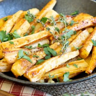 A close up of honey roast parsnips with golden crispy crusts, garnished with parsley and thyme.