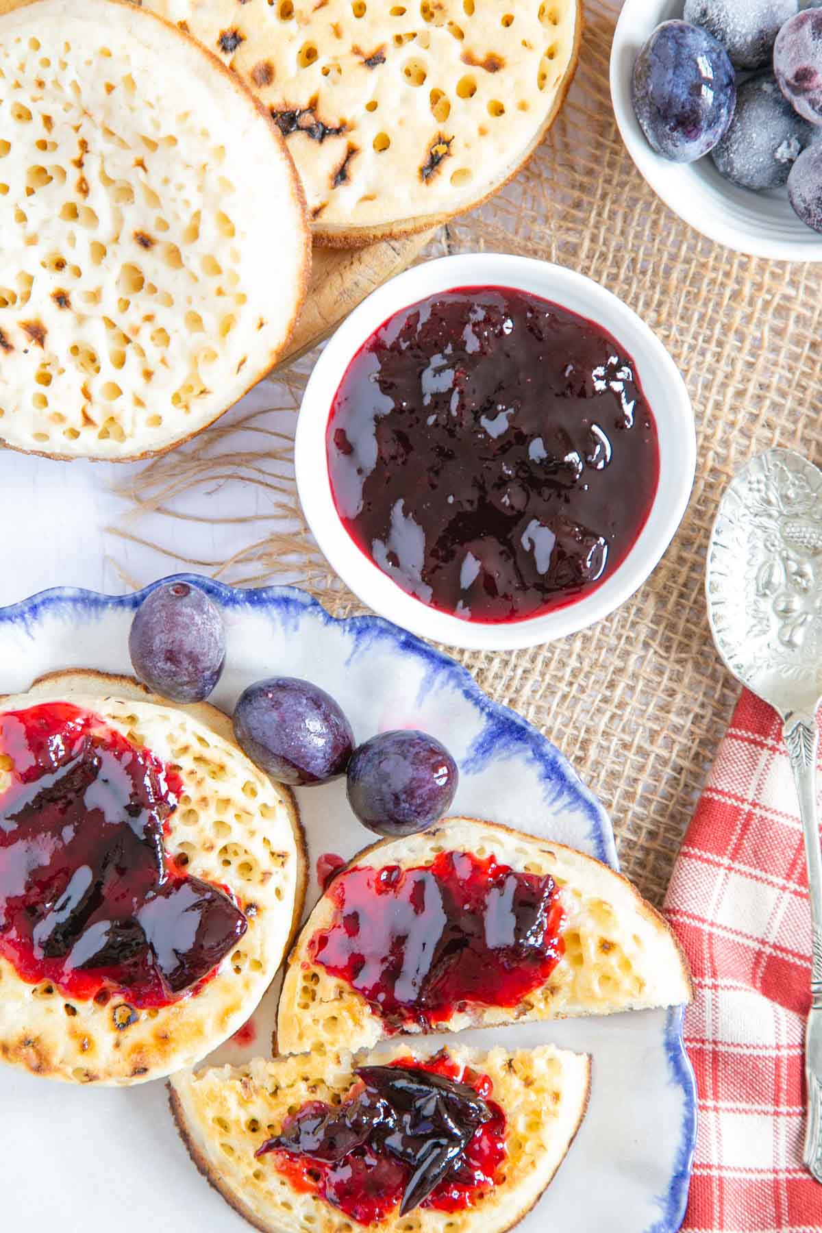 A toasted crumpet spread with butter and damson jam, with more in a small dish next to the plate.