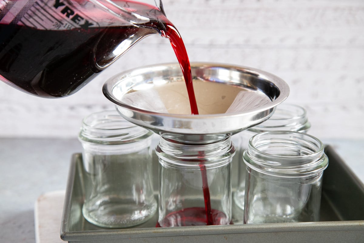 The jelly is being poured into pre-heated jars using a heatproof jug and a jam funnel.