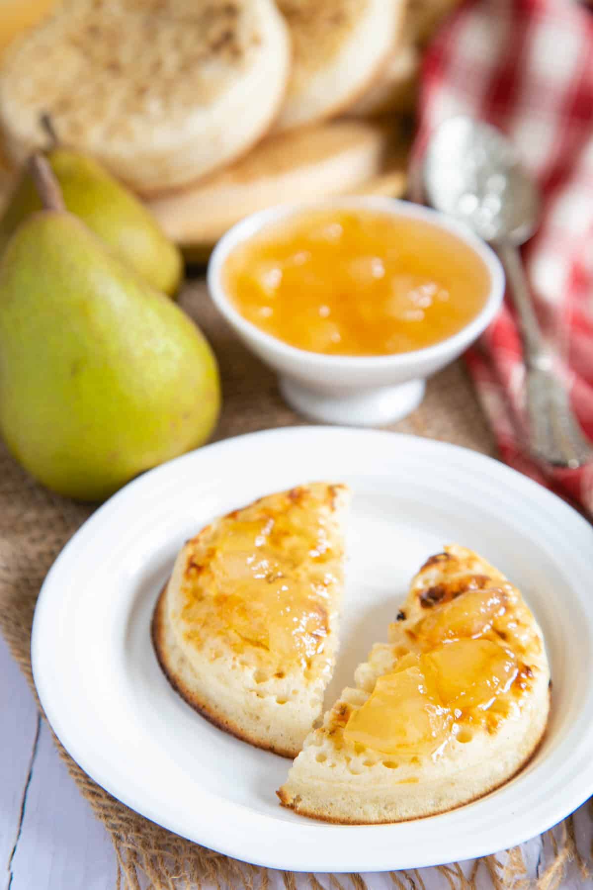A toasted crumpet spread with butter and golden pear jam. More jam in a small dish and more crumpets in the background.