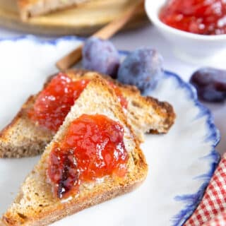 Toast spread with plum and apple jam, set on a white plate with a blue rim, contrating with a red gingham cloth.
