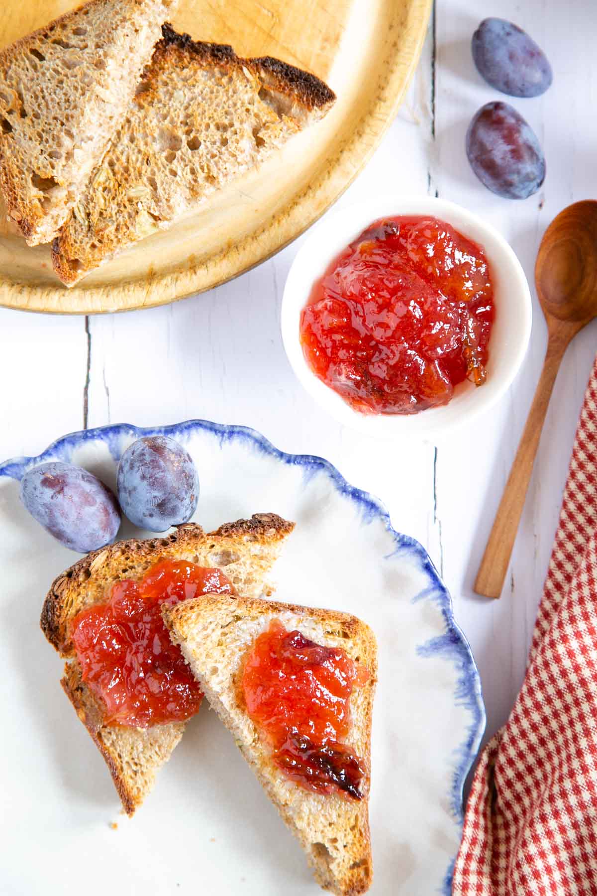 On a rustic whitewashed table, a blue and white plate of toast spread with plum and apple jam, a dish of the jam and fresh toast on a wooden cutting board.