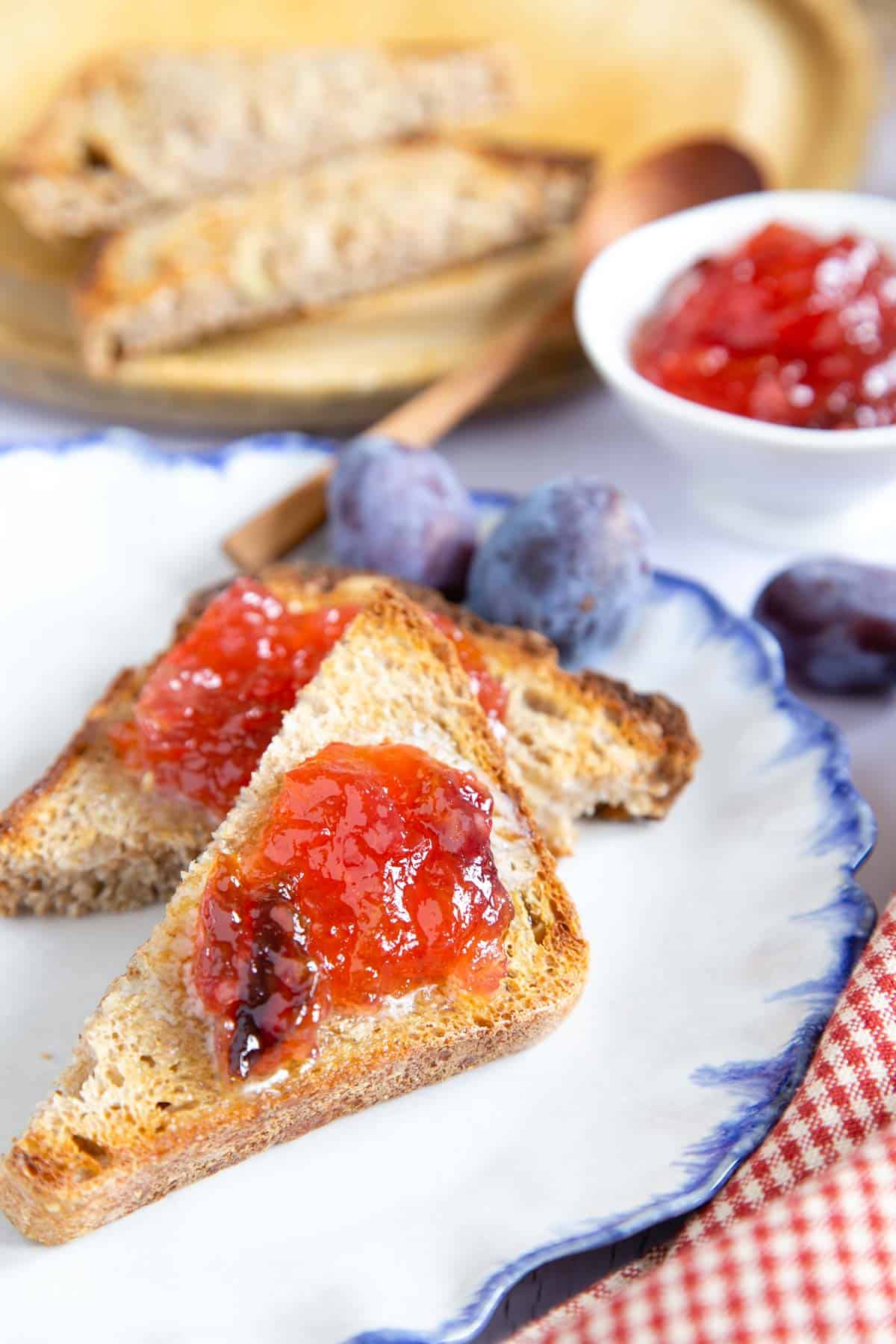Toast spread with plum and apple jam, set on a white plate with a blue rim, contrating with a red gingham cloth.