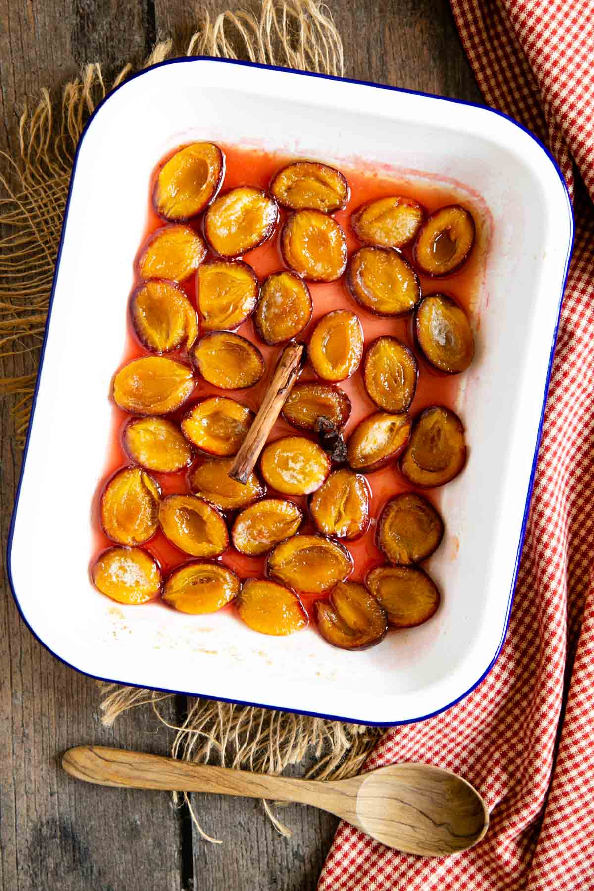 A white enamel roasting tin full of baked plums, sitting in a delicate pink syrup.