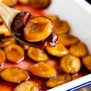 A close up of a soft roast plum, in a wooden spoon, served from a dish full of more plums.
