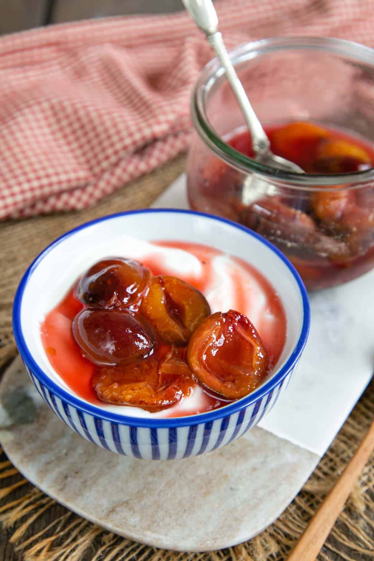 Stewed plums in their juice, served on a helping of yoghurt. More cooked plums are in a jar in the background.