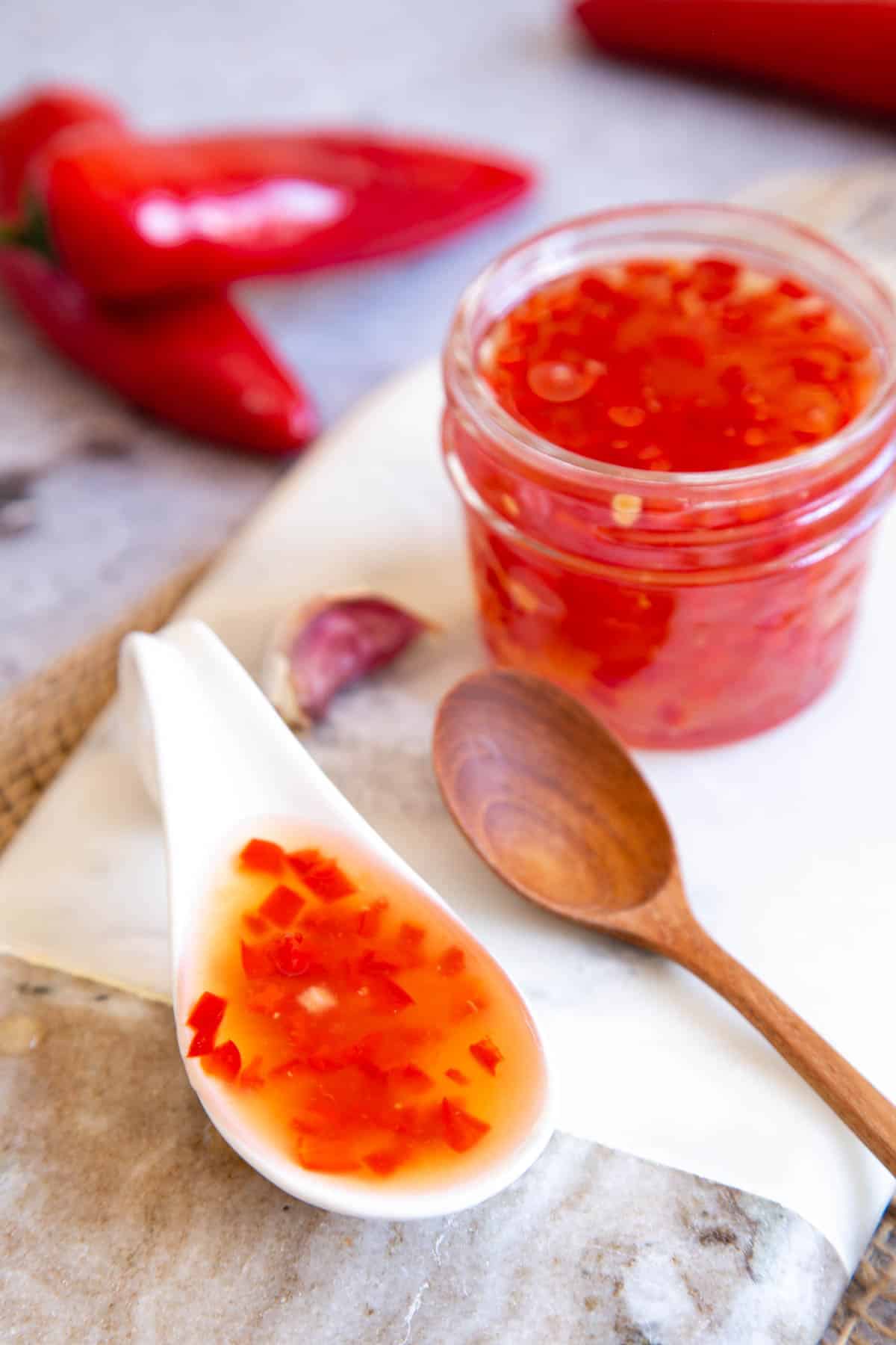 A jar of sweet chilli sauce with a wooden spoon, and an Asian style ceramic spoon full of the sauce. The warm red of the sauce contrasts with the cool white and grey marrble of the serving board on which it stands.