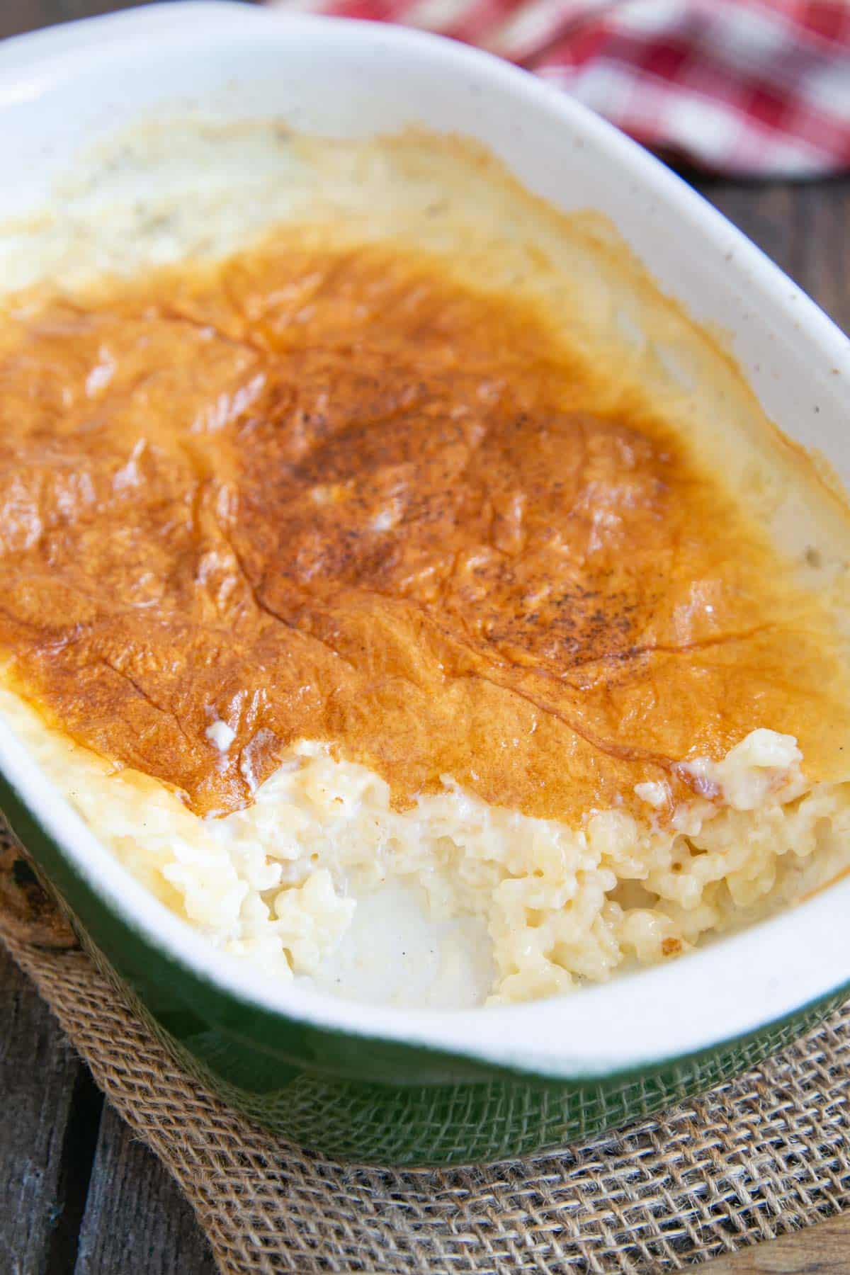A delicious baked rice pudding, in a white and green serving dish, with one helping removed. The soft crust on the pudding is golden brown.