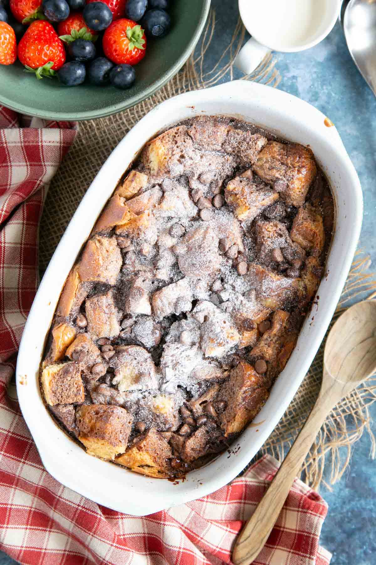A dish of chocolate hazelnut bread and butter pudding, dusted with icing sugar with a bowl of berries on the side.